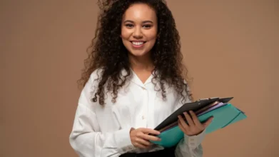 Processos Seletivos 5 Mulher sorridente com cabelos cacheados, vestindo camisa branca, segurando pastas e prancheta em frente a fundo bege. Foto: Freepik