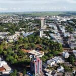 Imagem aérea da cidade de Cafelândia, no estado do Paraná, mostrando uma paisagem urbana bem organizada com prédios residenciais e comerciais cercados por ruas arborizadas. Ao fundo, é possível ver áreas industriais e extensas plantações que indicam a força do setor agrícola na região. A cidade apresenta um ambiente limpo, com boa infraestrutura e construções de diferentes tamanhos integradas à natureza local.