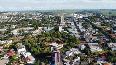 Imagem aérea da cidade de Cafelândia, no estado do Paraná, mostrando uma paisagem urbana bem organizada com prédios residenciais e comerciais cercados por ruas arborizadas. Ao fundo, é possível ver áreas industriais e extensas plantações que indicam a força do setor agrícola na região. A cidade apresenta um ambiente limpo, com boa infraestrutura e construções de diferentes tamanhos integradas à natureza local.
