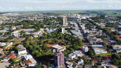 Imagem aérea da cidade de Cafelândia, no estado do Paraná, mostrando uma paisagem urbana bem organizada com prédios residenciais e comerciais cercados por ruas arborizadas. Ao fundo, é possível ver áreas industriais e extensas plantações que indicam a força do setor agrícola na região. A cidade apresenta um ambiente limpo, com boa infraestrutura e construções de diferentes tamanhos integradas à natureza local.