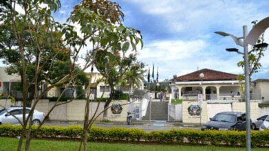 Imagem da fachada principal da Prefeitura Municipal de Cajamar, localizada no estado de São Paulo. A entrada é composta por uma escadaria central ladeada por portões brancos com o brasão da cidade estampado. Ao fundo, há um prédio de telhado cerâmico com colunas brancas e uma área coberta, onde algumas pessoas circulam. Árvores e arbustos bem cuidados emolduram a cena, e bandeiras oficiais estão hasteadas. A imagem transmite uma atmosfera organizada e acolhedora do espaço público municipal.