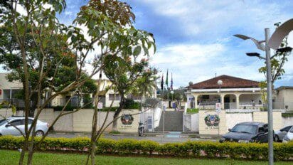 Imagem da fachada principal da Prefeitura Municipal de Cajamar, localizada no estado de São Paulo. A entrada é composta por uma escadaria central ladeada por portões brancos com o brasão da cidade estampado. Ao fundo, há um prédio de telhado cerâmico com colunas brancas e uma área coberta, onde algumas pessoas circulam. Árvores e arbustos bem cuidados emolduram a cena, e bandeiras oficiais estão hasteadas. A imagem transmite uma atmosfera organizada e acolhedora do espaço público municipal.