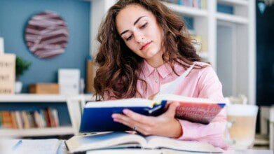 Mulher com cabelo castanho ondulado estuda em uma biblioteca, vestindo camisa rosa e lendo um livro com outros livros abertos à sua frente. Foto: Freepik