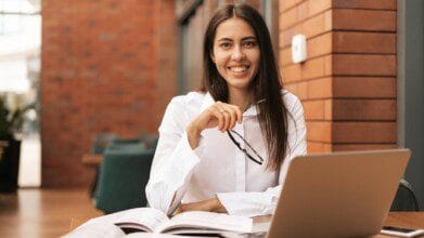 Mulher sorridente sentada à mesa com notebook, livros abertos e óculos na mão, em ambiente interno com parede de tijolos ao fundo.