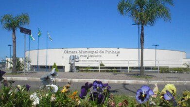 Fachada da Câmara Municipal de Pinhais com jardim florido em primeiro plano, palmeiras e bandeiras hasteadas ao fundo. Foto: SamirNosteb/Wikipédia