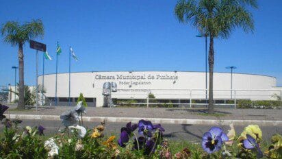 Fachada da Câmara Municipal de Pinhais com jardim florido em primeiro plano, palmeiras e bandeiras hasteadas ao fundo. Foto: SamirNosteb/Wikipédia