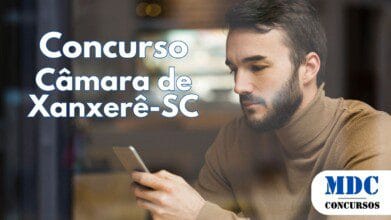 Homem jovem com barba e suéter marrom sentado em um café, olhando para o celular com expressão concentrada. A cena é vista através de um vidro, refletindo o ambiente ao redor, que inclui luzes desfocadas e móveis modernos no fundo - Câmara de Xanxerê (SC) oferece salários de Até R$ 5.495 em Concurso