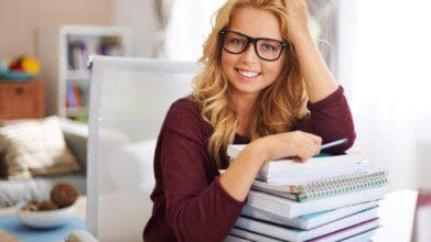 Mulher jovem sorridente com óculos e blusa vinho sentada à mesa, apoiando-se em uma pilha de livros e cadernos, segurando uma caneta.