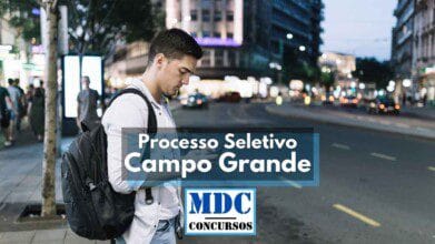 Processos Seletivos 4 A imagem mostra um jovem de mochila nas costas e camisa branca olhando para o celular enquanto caminha em uma avenida urbana ao entardecer. Ao fundo, há prédios iluminados, carros em movimento e pessoas caminhando pela calçada. No centro da imagem, aparece um banner com os dizeres “Processo Seletivo Campo Grande” e abaixo o logotipo da MDC Concursos.