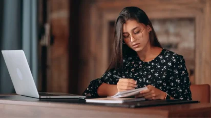 Mulher com óculos escrevendo em caderno enquanto usa notebook sobre a mesa. Foto: teksomolika/Freepik