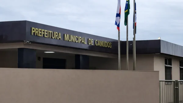 Fachada da Prefeitura Municipal de Canudos com letreiro em destaque e mastros com bandeiras em frente ao prédio sob céu nublado.