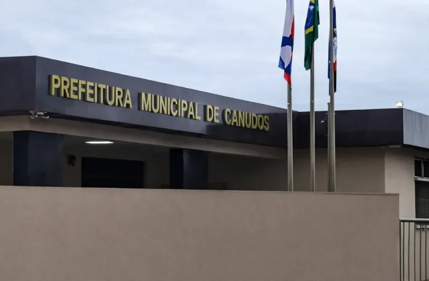 Fachada da Prefeitura Municipal de Canudos com letreiro em destaque e mastros com bandeiras em frente ao prédio sob céu nublado.