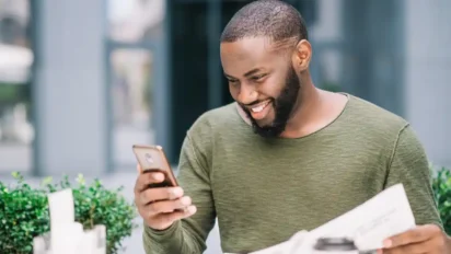 Homem sorridente segurando celular e jornal enquanto está sentado ao ar livre. Foto: Freepik