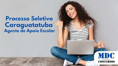 Mulher jovem de cabelos cacheados e sorriso alegre sentada no chão com as pernas cruzadas, usando jeans, camiseta listrada e tênis branco. Ela segura um laptop no colo e aponta com o polegar para o lado, expressando entusiasmo. O fundo é uma parede cinza lisa, e o chão tem aparência de madeira - Caraguatatuba (SP) oferece 232 vagas na Educação