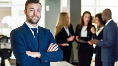 Homem de terno azul sorrindo com braços cruzados em escritório, enquanto pessoas ao fundo conversam em grupo. Foto: javi_indy/Freepik