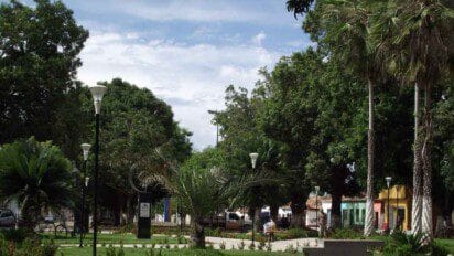 Praça arborizada em Carolina-MA com postes de luz, jardins bem cuidados, palmeiras, árvores grandes e casas coloridas ao fundo. Foto: Fernando Santos Cunha Filho/Wikipédia