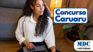 Mulher jovem de cabelos cacheados e pele morena sentada no chão com um laptop no colo, usando uma camisa branca e olhando pensativa para o lado. O ambiente tem iluminação aconchegante, com um abajur aceso sobre uma mesa de madeira ao lado de um sofá cinza e uma planta verde em um vaso trançado - Caruaru (PE) abre 29 vagas com salários de até R$ 5 mil em Concurso Público