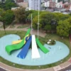 Vista aérea de rotatória urbana em Cascavel, com monumento central nas cores da bandeira do Brasil, fontes de água, áreas verdes arborizadas, ciclovia ao redor e prédios ao fundo. Foto: Divulgação/Secom – Prefeitura Municipal de Cascavel