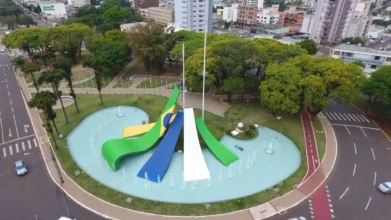 Cascavel/PR lança processo seletivo com 50 vagas para Professor 2 Vista aérea de rotatória urbana em Cascavel, com monumento central nas cores da bandeira do Brasil, fontes de água, áreas verdes arborizadas, ciclovia ao redor e prédios ao fundo. Foto: Divulgação/Secom – Prefeitura Municipal de Cascavel