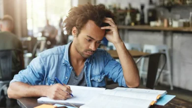 Homem estudando concentrado em mesa, escrevendo em caderno aberto enquanto apoia a cabeça com a mão em ambiente interno com outras pessoas ao fundo. Foto: wayhomestudio/Freepik