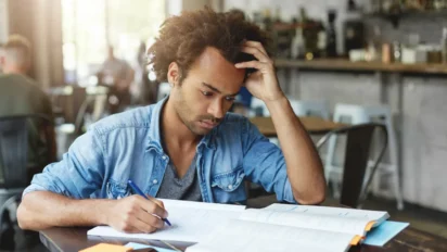 Homem estudando concentrado em mesa, escrevendo em caderno aberto enquanto apoia a cabeça com a mão em ambiente interno com outras pessoas ao fundo. Foto: wayhomestudio/Freepik