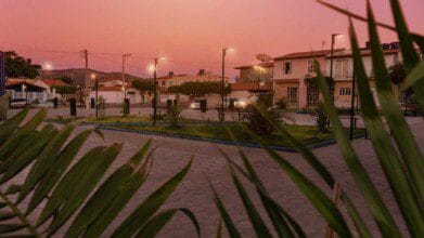 Praça iluminada ao entardecer em Caturama (BA), com bancos, postes, vegetação e casas ao fundo, vista através de folhas em primeiro plano. Foto: Divulgação/Prefeitura de Caturama-BA