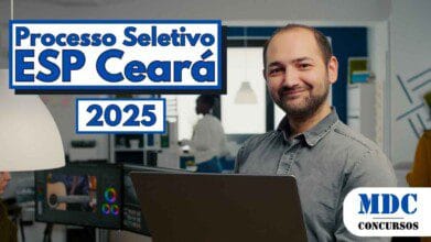 Processos Seletivos 4 Homem sorridente com barba rala e cabelo curto segura um notebook prateado em um escritório moderno e bem iluminado, usando camisa cinza de botão. Ao fundo, outras pessoas trabalham em mesas com computadores e prateleiras com materiais organizados. A decoração do ambiente é contemporânea, com paredes em azul e branco, luminárias pendentes e janelas grandes que permitem entrada de luz natural - Ceará abre 7 Vagas + CR com salários de até R$ 7 mil em Processo Seletivo na ESP/CE