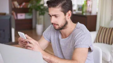 Homem sentado em frente a um notebook, segurando um celular e olhando atentamente para a tela, em ambiente interno com decoração ao fundo.