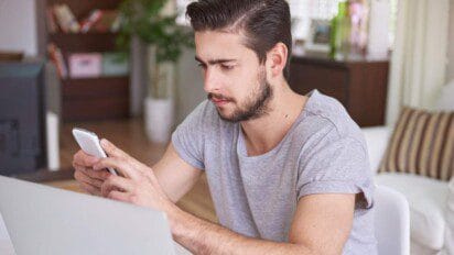 Homem sentado em frente a um notebook, segurando um celular e olhando atentamente para a tela, em ambiente interno com decoração ao fundo.