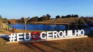 Letreiro com a frase "#EU❤️CERQUILHO" em frente a um lago, com palmeiras e pessoas caminhando ao fundo.