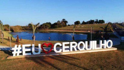 Letreiro com a frase "#EU❤️CERQUILHO" em frente a um lago, com palmeiras e pessoas caminhando ao fundo.