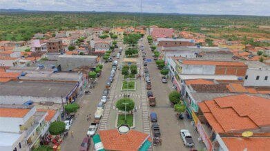 Vista aérea de Pintadas (BA) destaca avenida central arborizada, comércios, construções com telhados cerâmicos, áreas residenciais e vegetação do semiárido sob céu parcialmente nublado.