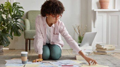 Como estudar após o trabalho: estratégias para quem tem pouco tempo 4 Como estudar após o trabalho estratégias para quem tem pouco tempo: Mulher de óculos vermelhos ajoelhada no chão, organizando papéis ao lado de um laptop aberto, com café e livros ao redor. Foto: wayhomestudio/Freepik