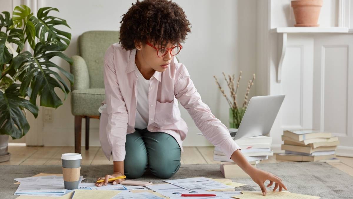 Como estudar após o trabalho: estratégias para quem tem pouco tempo 1 Como estudar após o trabalho estratégias para quem tem pouco tempo: Mulher de óculos vermelhos ajoelhada no chão, organizando papéis ao lado de um laptop aberto, com café e livros ao redor. Foto: wayhomestudio/Freepik