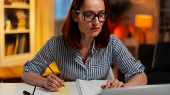 Como estudar legislação seca: Mulher com cabelos ruivos, óculos e blusa listrada estuda à noite em uma mesa, escrevendo em um caderno com post-its e usando um laptop. Foto: Freepik