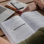 Como manter a motivação durante a preparação para concursos: Pessoa estudando com apostila aberta, notebook ao lado e livros empilhados sobre a mesa. Foto: Avery Evans/Unsplash