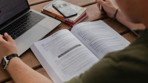 Como manter a motivação durante a preparação para concursos: Pessoa estudando com apostila aberta, notebook ao lado e livros empilhados sobre a mesa. Foto: Avery Evans/Unsplash