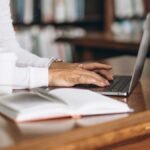 Uma pessoa sentada em uma mesa de madeira, digitando em um laptop ao lado de uma pilha de livros e uma xícara de café. Foto: senivpetro/Freepik