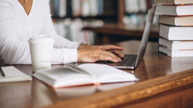 Uma pessoa sentada em uma mesa de madeira, digitando em um laptop ao lado de uma pilha de livros e uma xícara de café. Foto: senivpetro/Freepik