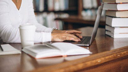 Uma pessoa sentada em uma mesa de madeira, digitando em um laptop ao lado de uma pilha de livros e uma xícara de café. Foto: senivpetro/Freepik
