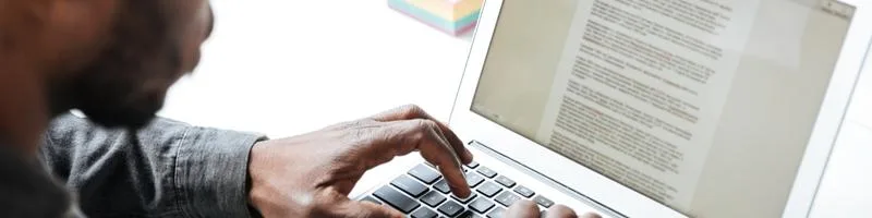 UFBA abre processo seletivo para Assistente Pedagógico 2 Pessoa digitando em um laptop, com foco nas mãos no teclado e na tela exibindo um texto.