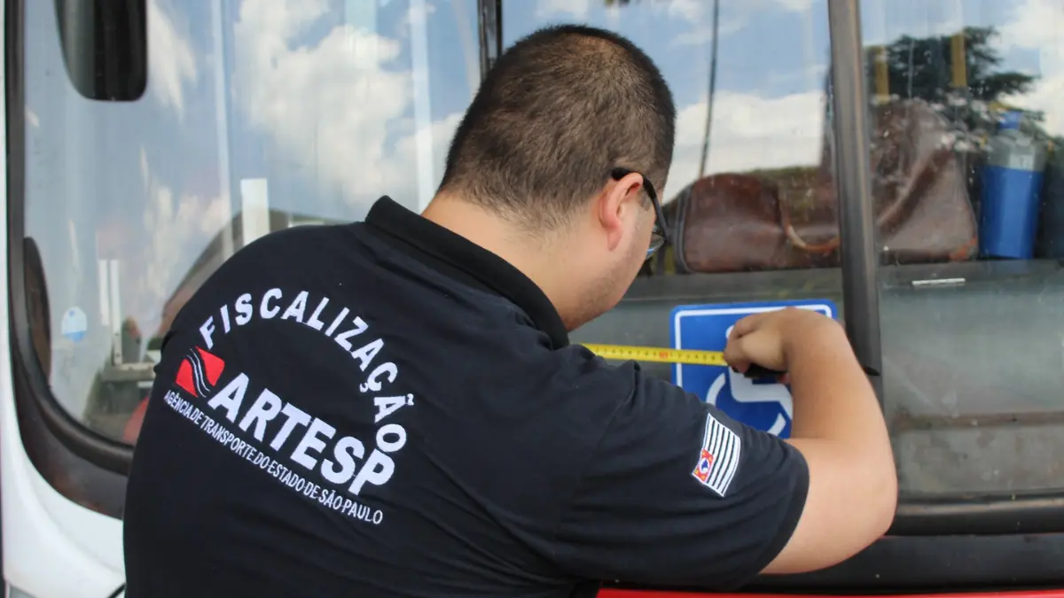 Agente de fiscalização do concurso ARTESP de costas medindo adesivo em porta de ônibus