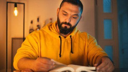 Homem com barba e moletom amarelo estuda concentrado à mesa, folheando um livro sob luz ambiente.