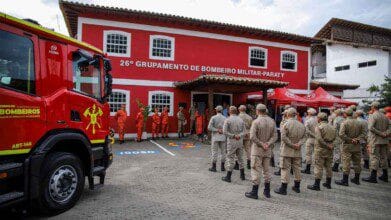 Imagem mostra uma cerimônia em frente ao prédio do 26º Grupamento de Bombeiro Militar de Paraty, no estado do Rio de Janeiro. Bombeiros uniformizados estão perfilados, enquanto outros, de uniforme laranja, posicionam-se próximos à entrada do prédio vermelho com janelas brancas. À esquerda, há um caminhão do Corpo de Bombeiros - CBMERJ, e à direita, tendas vermelhas com a logomarca da corporação.