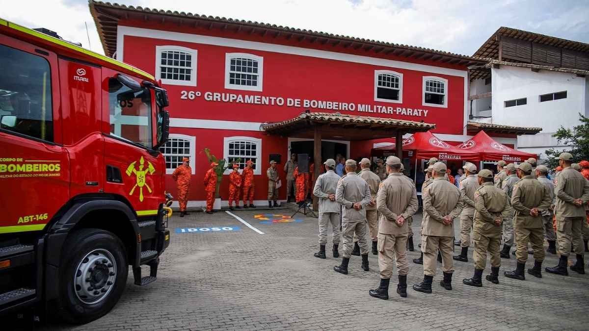 Concurso CBMERJ oferece 144 vagas para Soldado BM QBMP 1 Imagem mostra uma cerimônia em frente ao prédio do 26º Grupamento de Bombeiro Militar de Paraty, no estado do Rio de Janeiro. Bombeiros uniformizados estão perfilados, enquanto outros, de uniforme laranja, posicionam-se próximos à entrada do prédio vermelho com janelas brancas. À esquerda, há um caminhão do Corpo de Bombeiros - CBMERJ, e à direita, tendas vermelhas com a logomarca da corporação.