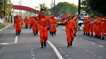 Bombeiros militares em uniforme laranja e banda desfilam em rua, concurso CBMMA