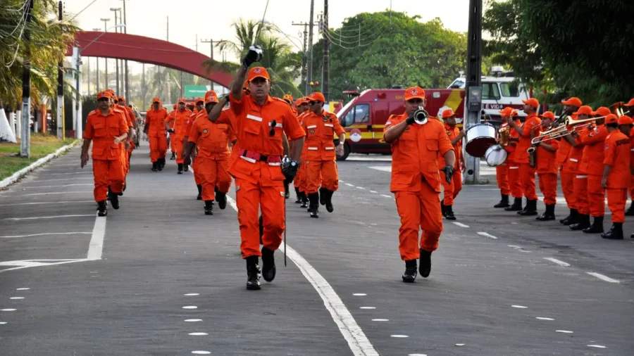 Bombeiros militares em uniforme laranja e banda desfilam em rua, concurso CBMMA