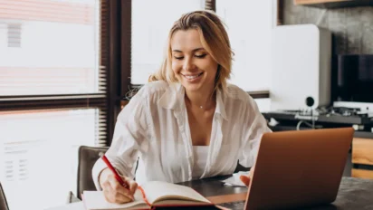 Mulher sorrindo enquanto estuda em uma mesa, escrevendo em caderno ao lado de um notebook em ambiente interno iluminado. Foto: senivpetro/Freepik
