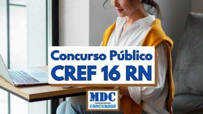 Mulher jovem de perfil leve sentada em ambiente interno iluminado, vestindo camisa branca com suéter mostarda sobre os ombros, digitando em notebook prateado apoiado em mesa de madeira clara próxima a janela; no centro da composição, banner retangular com fundo branco e bordas marrons exibe em letras azuis a frase “Concurso Público CREF 16 RN”; abaixo, logotipo quadrado da MDC Concursos em azul e verde sobre fundo branco