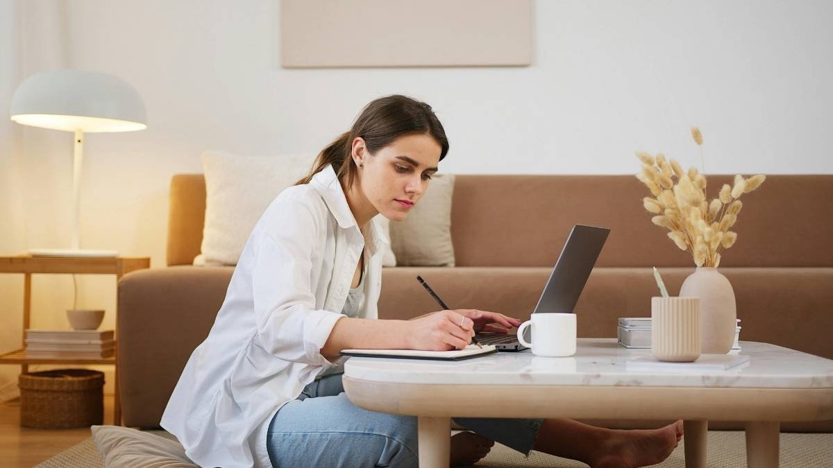 Mulher sentada no chão estudando com laptop e caderno sobre uma mesa de centro em ambiente doméstico.
